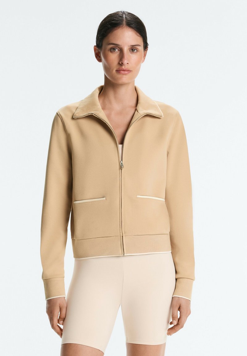 Femme portant une veste beige zippée avec des poches avant et un short de cycliste beige clair, debout devant un fond clair uni.