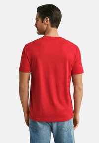 Mann mit dunklen Haaren, der ein einfarbiges rotes Kurzarm-T-Shirt und blaue Jeans trägt, von hinten vor einem schlichten weißen Hintergrund.