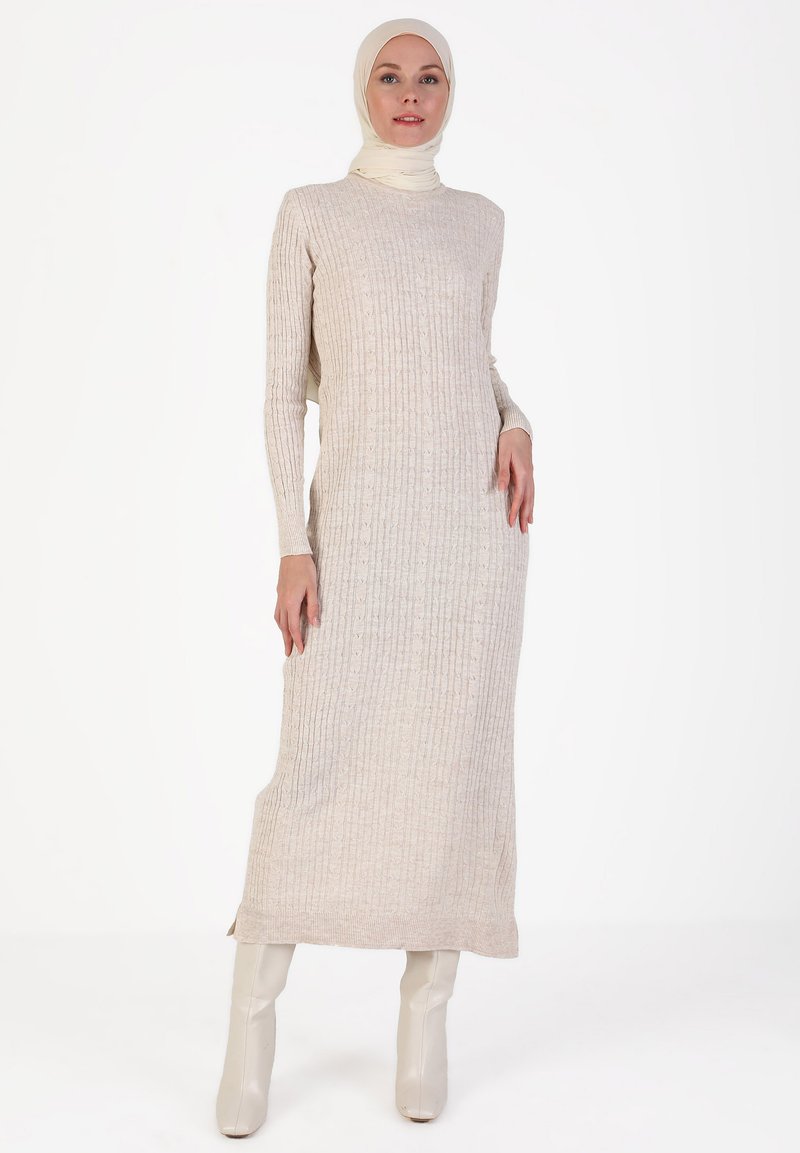 Robe en tricot côtelé beige à manches longues, col rond et fentes latérales. Portée avec des bottes à talons couleur crème, créant une silhouette élégante.