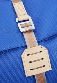 Gros plan sur un tissu bleu avec une sangle beige et une boucle argentée traversant un patch en cuir beige avec une sangle rayée orange.
