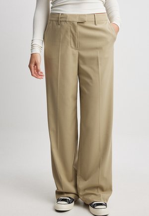 Persoon in beige wijde broek, witte langemouwen top en zwart-witte sneakers, met één hand in de zak.