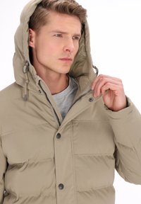 Beige-tikkitakki hupulla, edessä napit ja vetoketju. Ominaisuuksiin kuuluu vaakasuora tikkaukset ja pehmeä kangaspinta. Sisäkerroksena näkyy harmaa kerros.