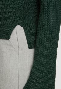 Strikket grønn genser med en teksturert, ribbet mønster, cropped design og lange ermer, kombinert med lysegråt stoff under.