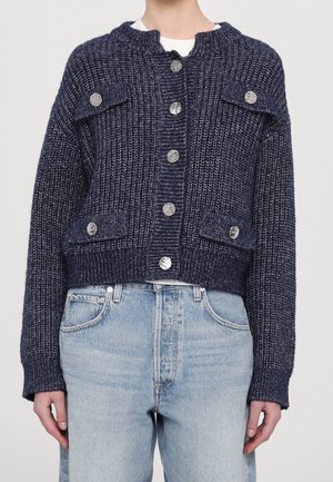 Personne portant un cardigan en maille texturée bleu foncé avec des boutons argentés et un jean taille haute bleu clair sur un fond uni.