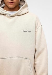 Beige hoodie gemaakt van zachte stof, met een voorzak en "CreateChange" tekst geborduurd op de linkerborst in donkere draad.