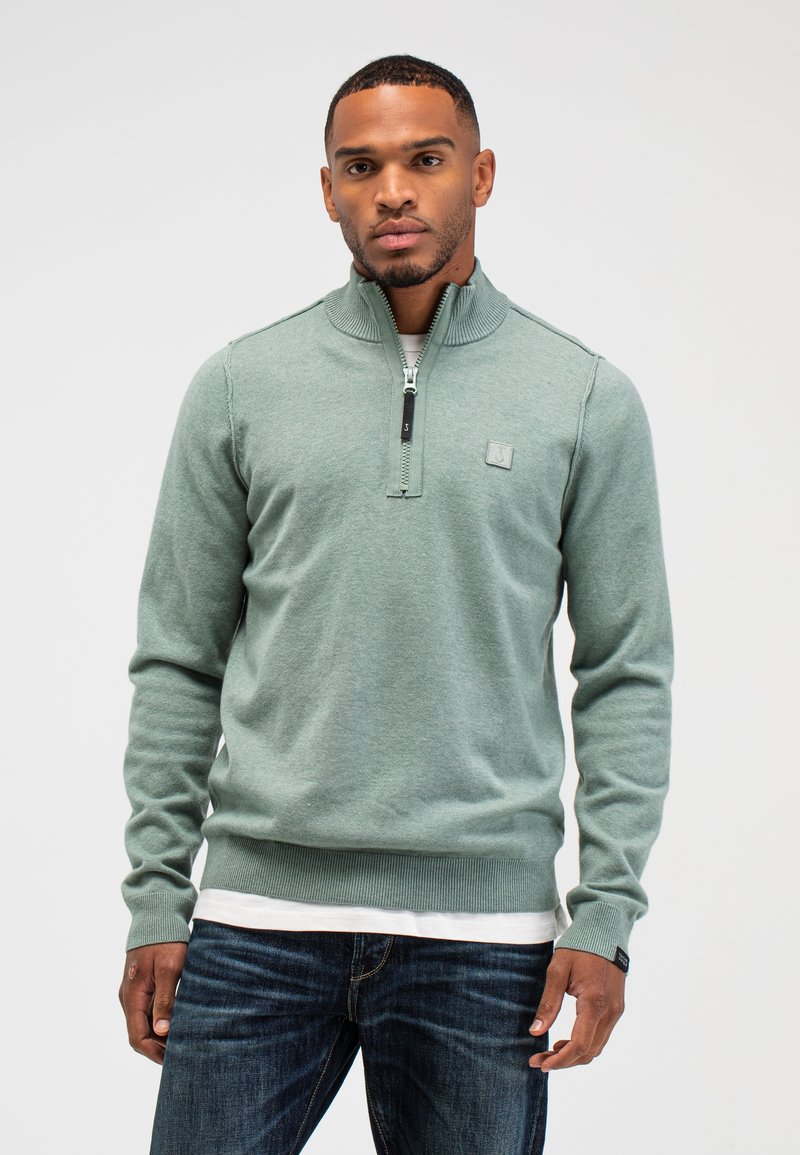 Lichtgroene zip-up trui met geribde kraag en manchetten, met een klein logopatch op de borst, gedragen over een wit shirt.