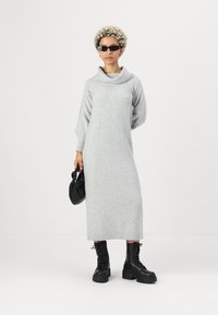 Vestido cinza de malha com gola alta e mangas longas, combinado com botas pretas robustas de atacadores e uma bolsa preta na mão.