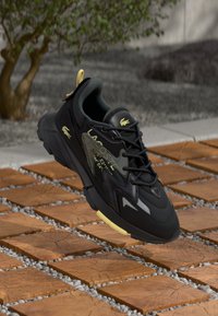 Black and yellow Lacoste sneaker floating above wooden tiles with small gray stones, outdoor setting with tree and concrete wall in background.