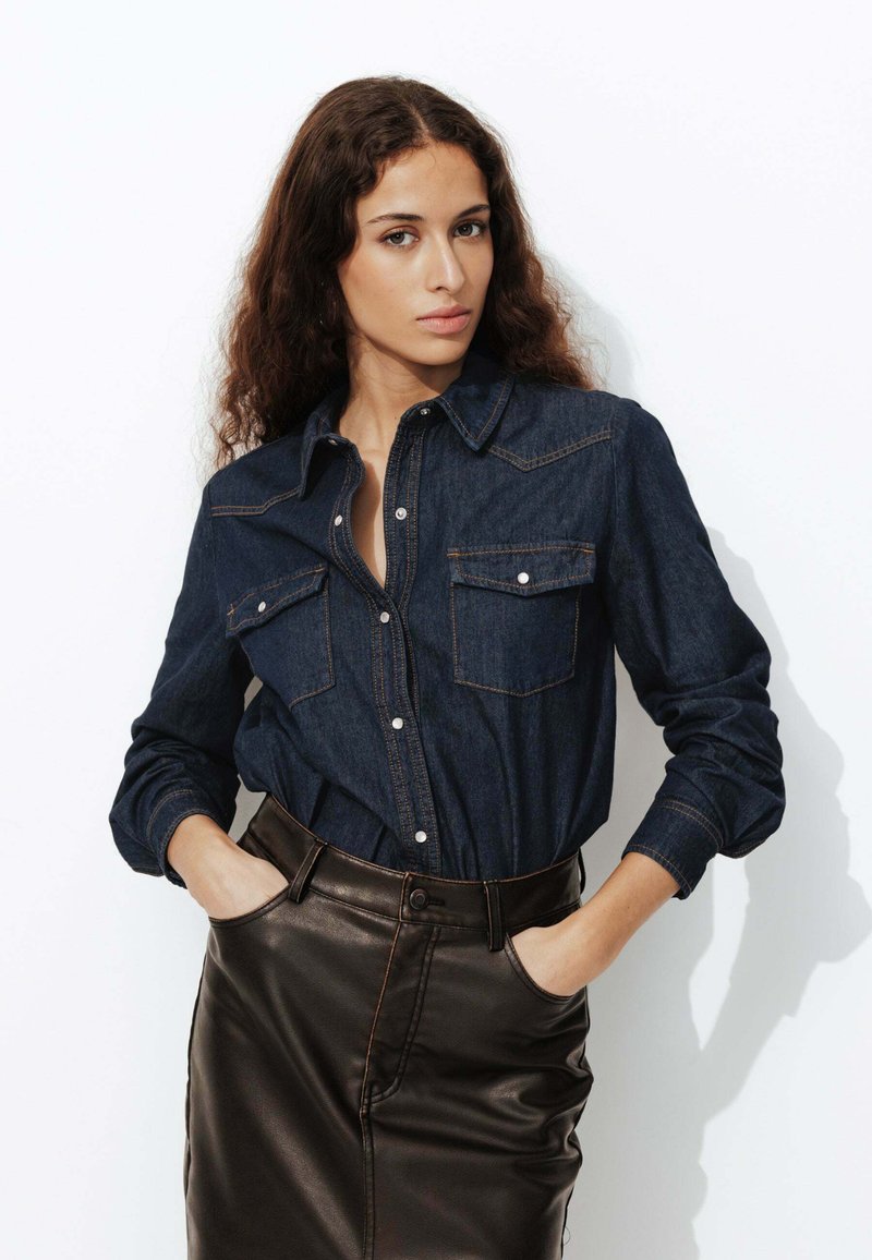 Chemise en denim bleu foncé à manches longues, avec deux poches poitrine et des coutures orange contrastantes. Associée à une jupe en cuir noire.