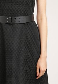 Gros plan sur une robe noire texturée avec une large ceinture en cuir noir, montrant la taille et le bras droit de la personne portant la robe sur un fond uni.