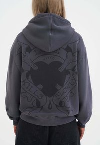 Felpa grigia con un grande grafico di un cuore circondato da cani, decorata con il testo "L'amore uccide lentamente". Presenta un cappuccio con stringhe.