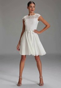Frau steht mit der Hand in der Hüfte, trägt ein ärmelloses weißes Spitzen- und Chiffon-Kleid in knielanger Form sowie silberne Riemchensandalen vor einem grauen Hintergrund.