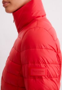 Czerwona pikowana kurtka z wysokim kołnierzem, z poziomym wzorem pikowania i naszywką z logo na lewym rękawie.