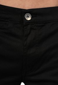 Un pantalon en denim noir possède un bouton métallique visible avec une marque, des détails cousus et une texture lisse. La ceinture est partiellement visible.