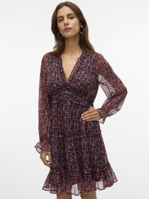 Frau trägt ein schwarzes und rotes Blumenkleid mit transparenten langen Ärmeln und einem gerafften Saum, posiert mit einer Hand in der Hüfte vor weißem Hintergrund.