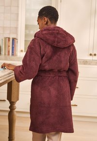 Accappatoio con cappuccio color bordeaux, in tessuto morbido e testurizzato, con cintura annodata in vita e maniche lunghe, visto da dietro.