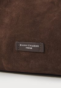 Bruine suède handtas met een leren merklabel met de gegraveerde tekst "GIANNI CHIARINI FIRENZE." Zachte textuur met een subtiele glans.