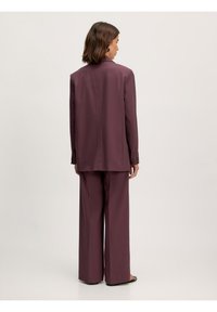 Dunkellila Anzug mit einem maßgeschneiderten Blazer und weiten Hosen. Der Stoff hat eine glatte Textur mit sichtbaren Nähten und minimalem Zubehör.