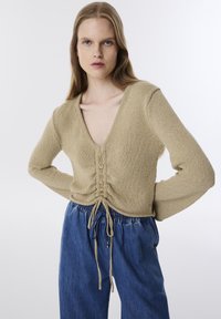 Beige gestrickter Pullover mit V-Ausschnitt, ausgestattet mit einem gerafften Design und Kordeln an der Taille. Akzentuiert mit Kordel-Details in der Mitte.