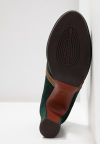 Semelle d'une botte à talon en daim vert avec une semelle intermédiaire en cuir marron, un talon en caoutchouc texturé, et le nom de la marque "OCHIE MIHARA" embossé sur la semelle.