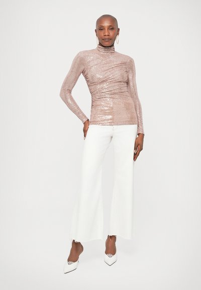 Λαμπερή ροζ χρυσή μπλούζα μακρύ μανίκι με ψηλό λαιμό και συγκεντρωμένη υφή, συνδυασμένη με λευκά τζιν καμπάνες και λευκά ψηλοτάκουνα παπούτσια.