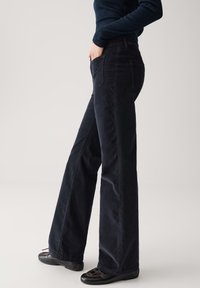 Persoon in hoge taille, wijde donkere ribfluwelen broeken met de handen in de zakken, gecombineerd met zwart platte schoenen en een donker geribbelde top.