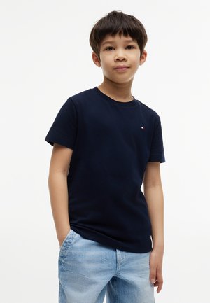 Dreng med kort mørkt hår iført en navyblå t-shirt og lyseblå jeans, står med den ene hånd i lommen mod en hvid baggrund.