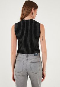 Čierny bezrukávový top s vertikálnymi bielymi prúžkami, skrátený strih. V kombinácii so šedými džínsami z denimu s zadnými vreckami a štítkom.