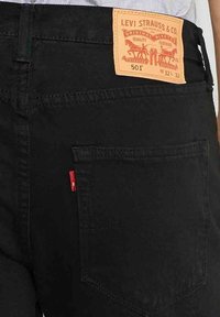 Zwarte denim jeans met een tan leren label met oranje tekst op de achtertailleband en een klein rood tabje op de achterzak.