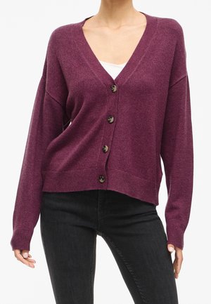 Femme portant un cardigan violet à col en V boutonné sur un haut blanc et un jean noir, debout, mains détendues le long du corps.