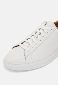 Λευκό δερμάτινο αθλητικό παπούτσι με στρογγυλή μύτη, επίπεδη καουτσούκ σόλα και παραδοσιακή λουρίδα κλεισίματος. Διαθέτει διακριτική σήμανση στο πλάι.