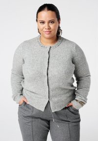 Vrouw draagt een grijze vest met knopen en kraalversieringen en grijze broek met krijtstrepen, staat met handen in de zakken tegen een witte achtergrond.