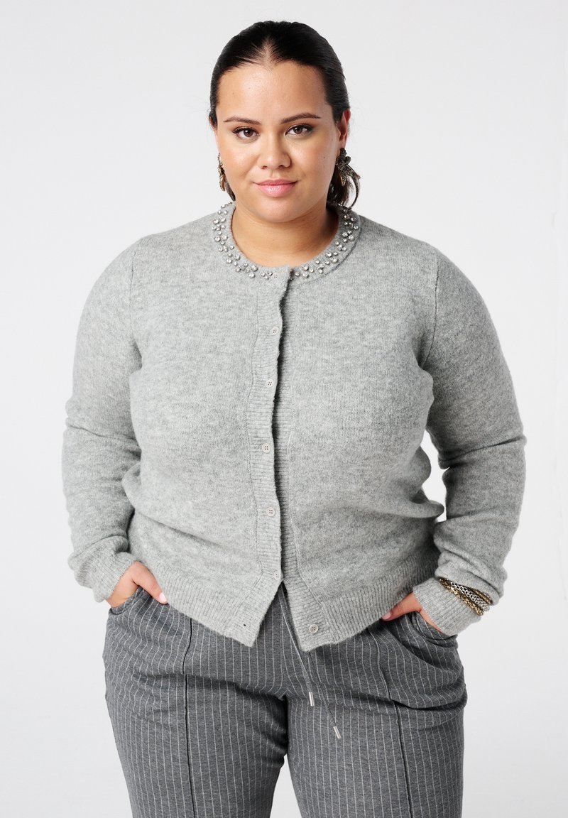 Vrouw draagt een grijze vest met knopen en kraalversieringen en grijze broek met krijtstrepen, staat met handen in de zakken tegen een witte achtergrond.