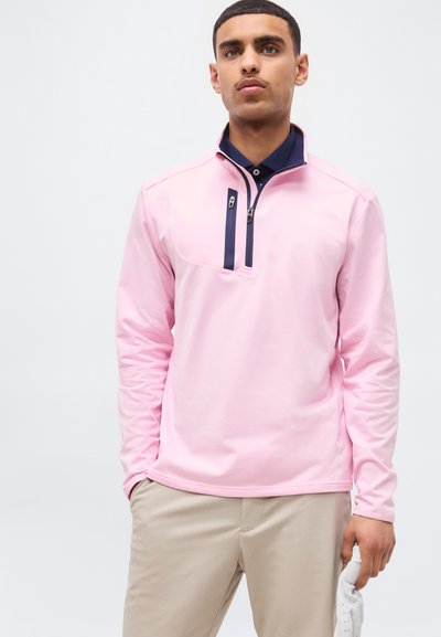 Młody mężczyzna w jasnoróżowym pulowerze z granatowymi detalami na zamku, beżowe spodnie, trzymający białą rękawiczkę golfową w prawej ręce.