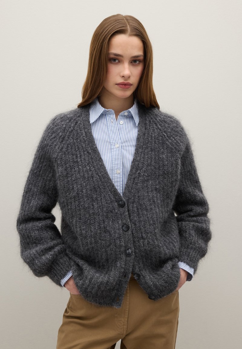 Cardigan lavorato a maglia grigio con finitura a rilievo, scollo a V e cinque bottoni scuri. Indossato sopra una camicia a righe bianche e blu.