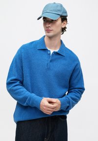 Jovem usando um boné azul claro e uma camisa polo azul de manga longa texturizada, com as mãos entrelaçadas à frente contra um fundo simples.