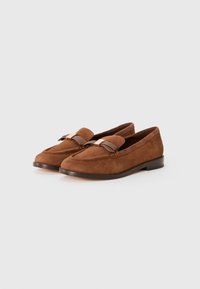 Brūnu zamša loafers pāris ar zemu papēdi un ādas izliektu detaļu augšpusē, uz viendabīga balta fona.