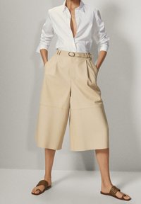 Beige wijde broek met plooien, met een aansluitende taille en een riem. Draagt een wit overhemd met knopen en bruine gevlochten sandalen.