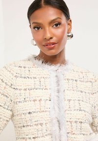 Balta teksturēta tweed jaka ar atvērtu priekšpusi, kurai ir maigs kažokādas apmalējums un smalki oranži un pelēki raksti. Spīdīgas sudraba gredzenveida auskari.