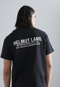 Must musta T-särk, millel seljal on suur valge tekst "HELMUT LANG" ning täiendav tekst, mis annab ülevaate limiteeritud väljaandest ja kauplustest.