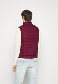 Burgundská prošívaná vesta s vysokým límcem, horizontální prošívaný design, lehký materiál, nošena přes texturovaný bílý svetr, doplněna o světlé džíny.