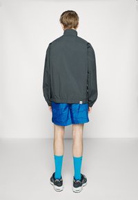 Grijze lichte jas met een hoge kraag, gecombineerd met blauw patroon shorts, blauwe sokken en zwarte sportschoenen, gezien vanaf de achterkant.
