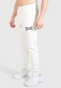 Vita sweatpants med svart "SMILODOX" text på vänster ben, ett elastiskt midjeband, sidofickor och ribbade muddar. Bärs med vita sneakers.