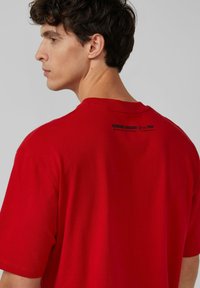 Rotes Baumwoll-T-Shirt mit kurzen Ärmeln, ausgestattet mit einem Rundhalsausschnitt und einem rückseitigen Grafiktext mit der Aufschrift "HANGING AROUND Seit 1950." Glatte Textur, lässiges Design.