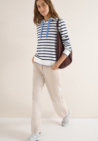 Jeune femme portant un sweat à capuche à rayures blanches et bleu marine avec des cordons bleus, un pantalon beige, des baskets argentées, et un sac à bandoulière marron.