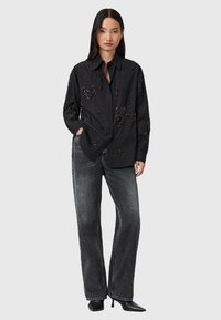 Donna che indossa una camicia nera a maniche lunghe con perline, jeans larghi neri sbiaditi e tacchi a punta neri, in piedi con una mano in tasca.