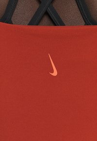 Reggiseno sportivo rosso elasticizzato con design a schiena scoperta nera. Caratterizzato da un piccolo logo arancione Nike sulla parte anteriore. Tessuto di consistenza liscia.