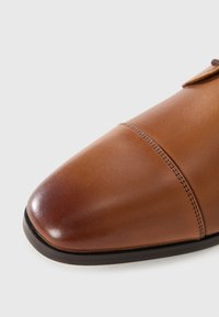 Zapato de cuero marrón con punta redondeada, textura suave y costuras decorativas en la parte superior. Suela de goma negra con acentos mínimos.