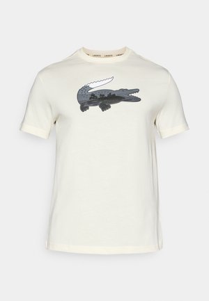 T-shirt in cotone color crema con un logo di coccodrillo grigio scuro posizionato in modo prominente al centro. Include un collo a giro e maniche corte.
