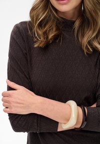 Frau mit hellbraunen Haaren, die ein dunkles, strukturiertes Langarmoberteil trägt und zwei auffällige Armbänder hat, eines in Creme und eines in dunkelbraun, auf ihren übergeschlagenen Armen.